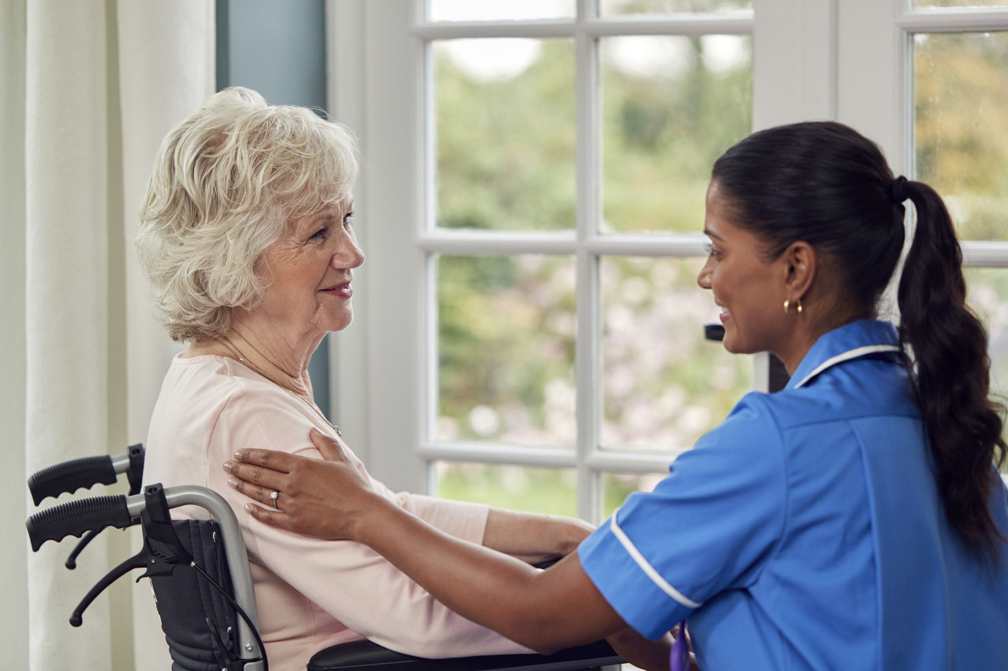 Female Care Worker In Uniform Talking With Senior Woman Sitting In Wheelchair In Care Home Lounge